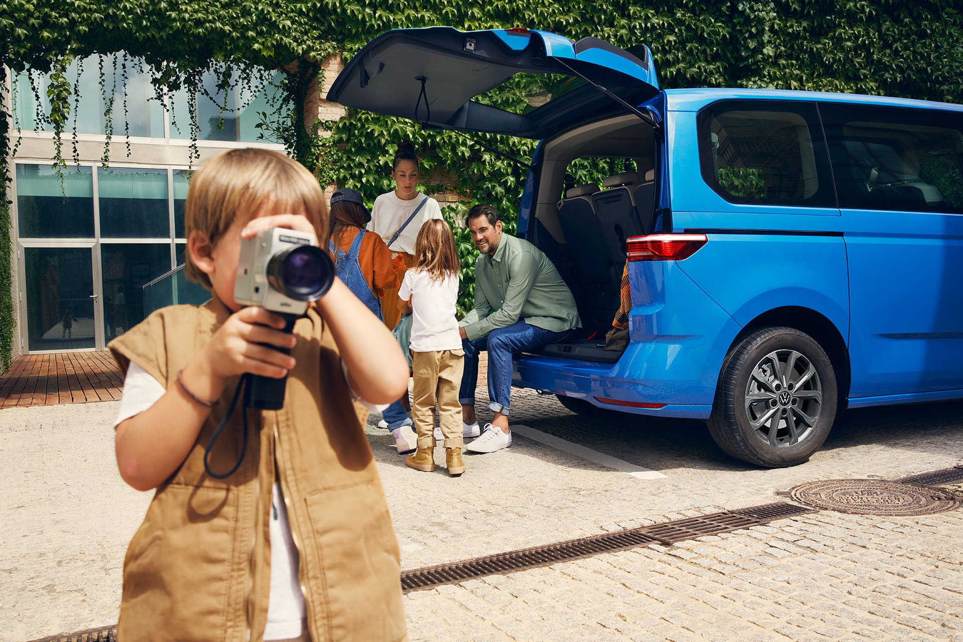 Chłopiec z małą przenośną kamerą na tle rodziny stojącej przy Volkswagenie Multivanie Family z otwartą klapą.